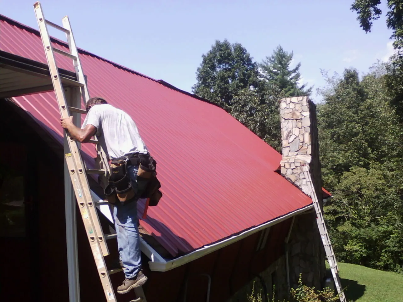 Roofer on a ladder beside a red metal roof and stone chimney — Soffit & Fascia Repair by Roofing Brothers in Miles